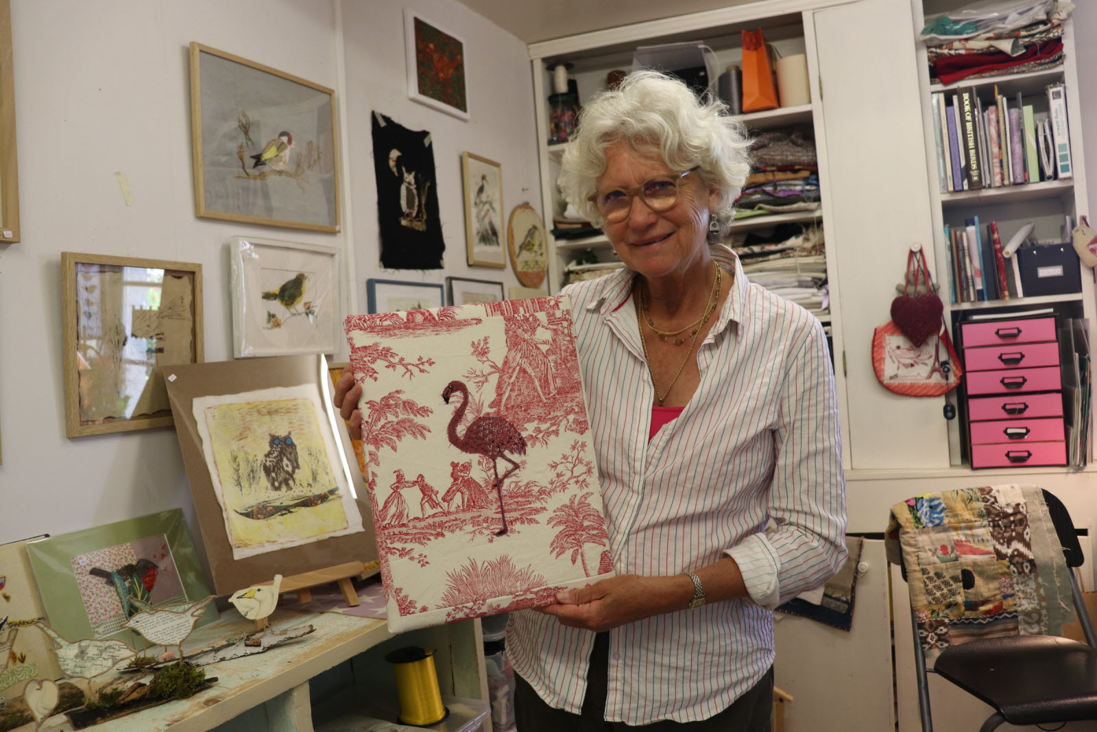 Une dame aux cheveux blancs tient un tableau en toile de jouy avec un flamand rose brodé